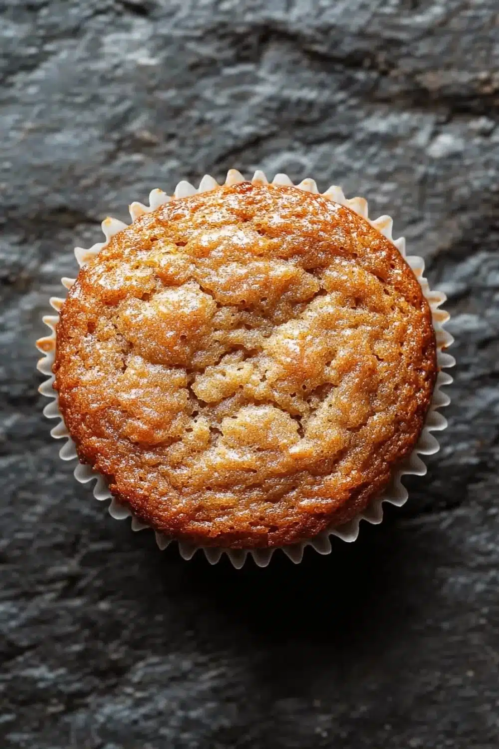 Banana muffin gluten free - the image is a close-up of a freshly baked muffin in a white paper liner. the muffin appears to be golden brown in color and has a crumbly texture. it is sitting on a dark, textured surface with a rough, uneven texture. the edges of the muffin are slightly curled, giving it a textured appearance. the background is blurred, making the muffins the focal point of the image.