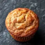 Banana muffin no added sugar - the image is a close-up of a muffin on a black textured surface. the muffin appears to be freshly baked and has a golden brown color. it has a crumbly texture on top, with small pieces of bread crumbs scattered throughout. the edges of the muffin are slightly curled, indicating that it has been freshly baked. the background is blurred, making the muffins the focal point of the image.