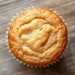 Banana muffin no added sugar - the image is a close-up of a freshly baked muffin. the muffin is golden brown in color and has a crumbly texture. it is sitting on a wooden surface with a textured surface. the top of the muffin has a swirl of powdered sugar on it, giving it a shiny appearance. the edges of the cupcake are lined with white paper liners. the background is blurred, making the muffins the focal point of the image.