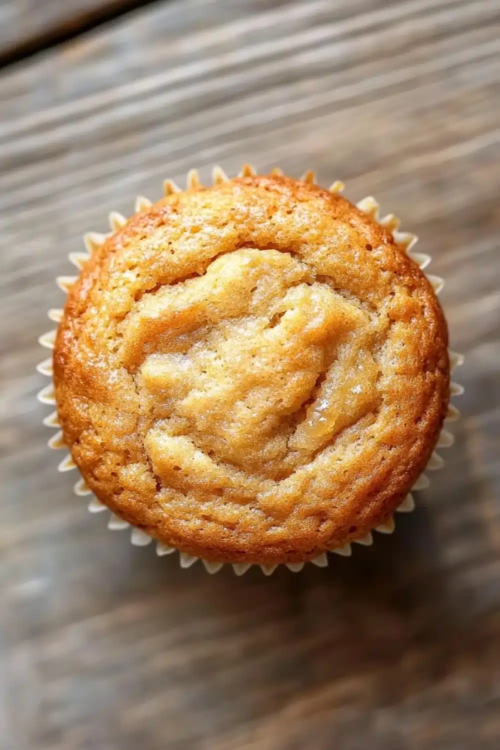 Banana muffin no added sugar - the image is a close-up of a freshly baked muffin. the muffin is golden brown in color and has a crumbly texture. it is sitting on a wooden surface with a textured surface. the top of the muffin has a swirl of powdered sugar on it, giving it a shiny appearance. the edges of the cupcake are lined with white paper liners. the background is blurred, making the muffins the focal point of the image.