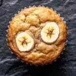 Banana muffin protein - the image is a close-up of a muffin in a white paper liner. the muffin appears to be freshly baked and has a golden brown color. on top of the muffin, there are two slices of banana arranged in a circular pattern. the banana slices are cut in half, revealing the soft, fluffy interior. the background is a dark, textured surface.