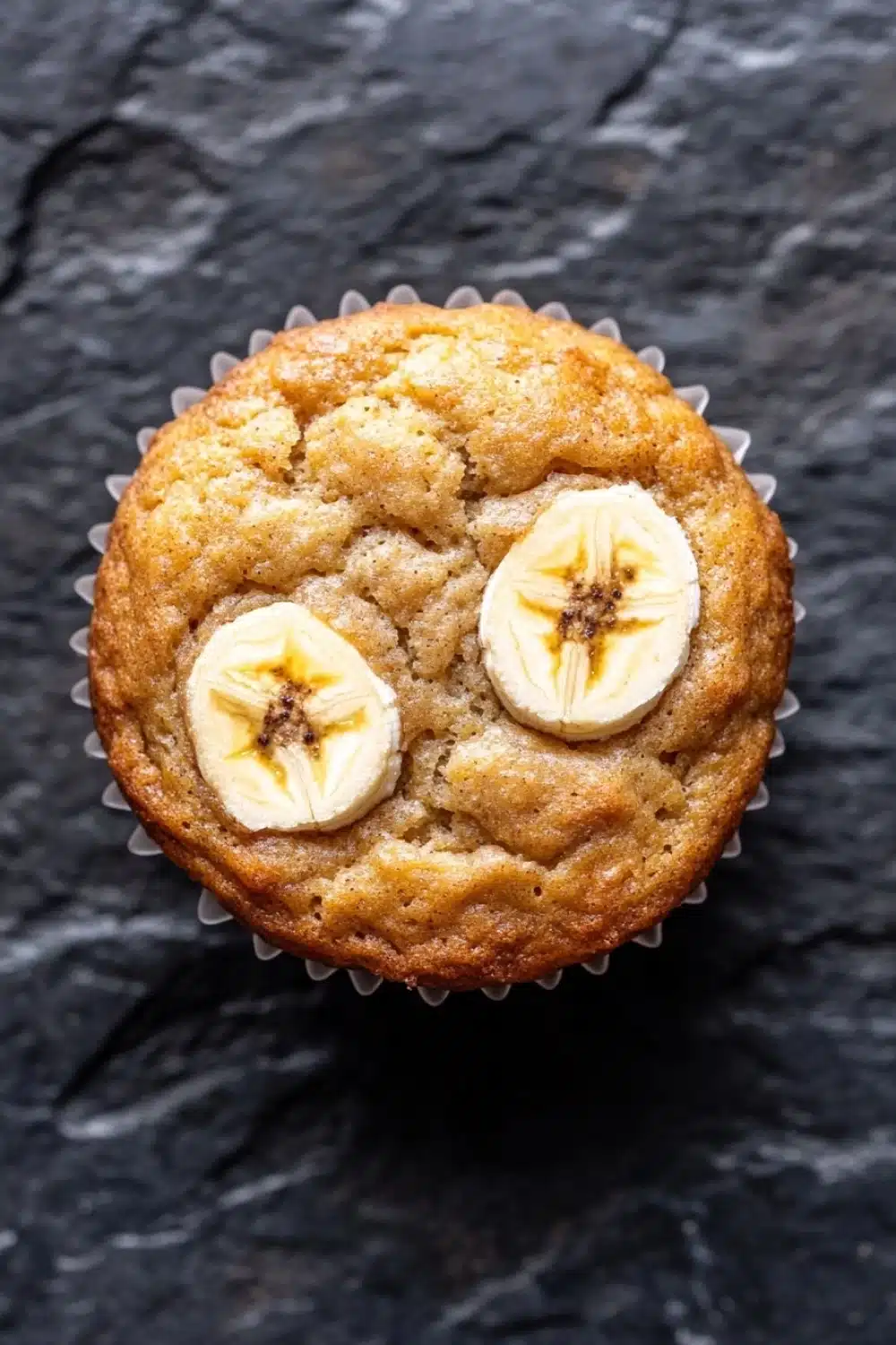 Banana muffin protein - the image is a close-up of a muffin in a white paper liner. the muffin appears to be freshly baked and has a golden brown color. on top of the muffin, there are two slices of banana arranged in a circular pattern. the banana slices are cut in half, revealing the soft, fluffy interior. the background is a dark, textured surface.