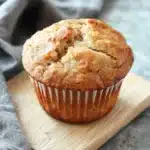 Banana muffin rustic bread - the image shows a freshly baked muffin on a wooden cutting board. the muffin is golden brown in color and has a crumbly texture. it is sitting on a gray countertop with a gray cloth napkin on the side. the background is blurred, but it appears to be a kitchen countertop.