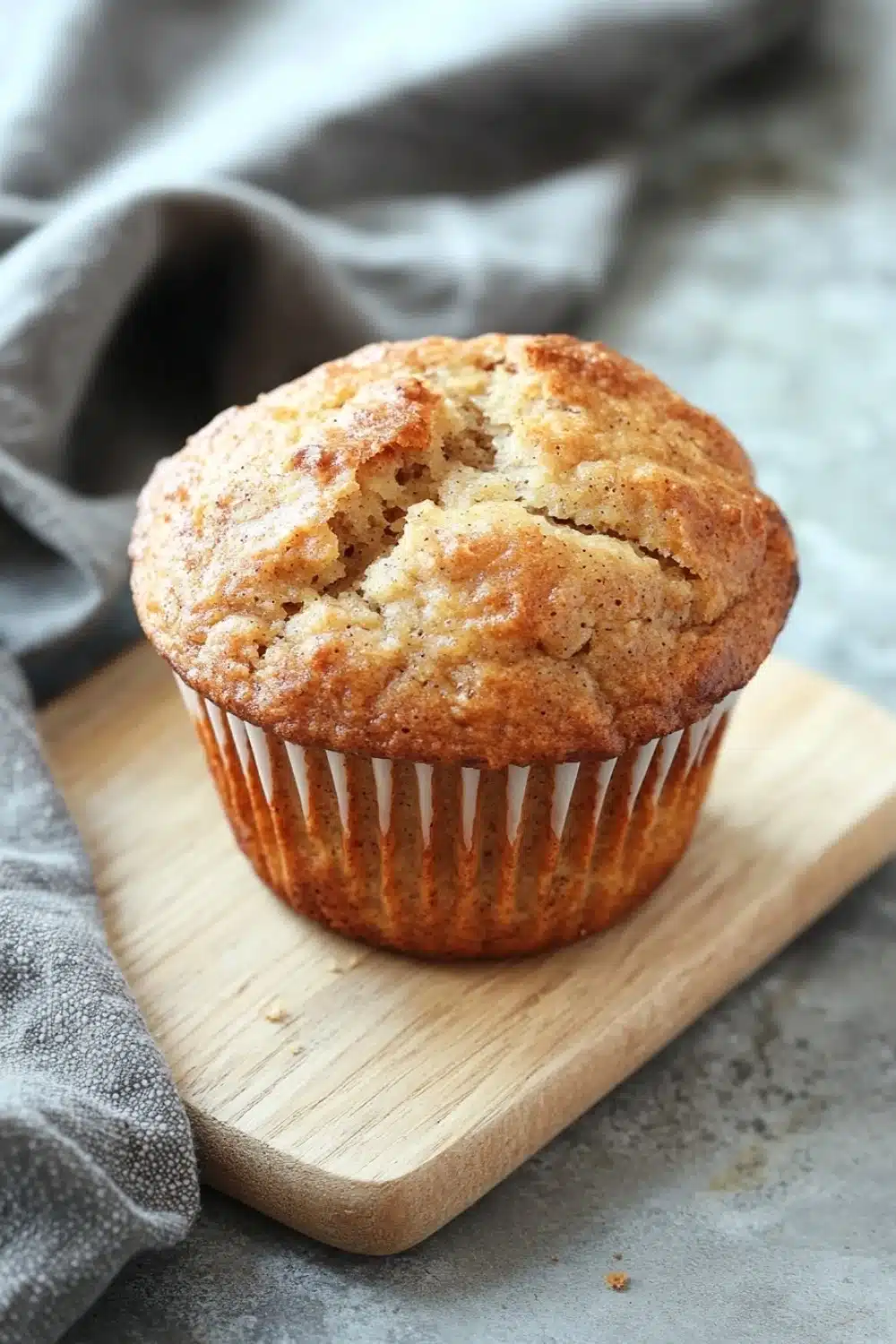 Banana muffin rustic bread - the image shows a freshly baked muffin on a wooden cutting board. the muffin is golden brown in color and has a crumbly texture. it is sitting on a gray countertop with a gray cloth napkin on the side. the background is blurred, but it appears to be a kitchen countertop.