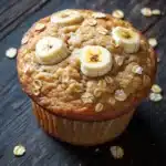 Banana oat yogurt muffin - the image is of a freshly baked banana muffin on a wooden surface. the muffin is golden brown in color and has a crumbly texture. on top of the muffin, there are several sliced bananas arranged in a circular pattern. the bananas are cut into thin slices and are scattered around the edges of the cupcake. the background is blurred, but it appears to be a dark wooden table. the overall mood of the image is warm and inviting.