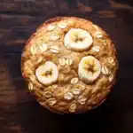 Banana oat yogurt muffin - the image is a close-up of a round, golden-brown muffin on a wooden surface. the muffin appears to be freshly baked and has a golden brown crust. on top of the muffin, there are three sliced bananas arranged in a circular pattern. the bananas are white and appear to be ripe and juicy. the background is dark and out of focus, making the muffins the focal point of the image.