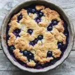 Blueberry muffin mix cobbler - the image is of a freshly baked blueberry pie in a round ceramic pie dish. the pie has a golden brown crumbly topping on top, with small blueberries scattered throughout. the blueberries are a deep purple color and appear to be ripe and juicy. the dish is sitting on a wooden surface, and the background is blurred, making the pie the focal point of the image.