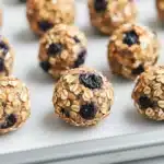 Blueberry muffin protein balls - the image shows a tray of freshly baked granola balls on a white marble countertop. the balls are round and golden brown in color, with visible oats and blueberries scattered throughout. they are arranged in a scattered manner, with some overlapping each other. the tray appears to be lined with parchment paper, and the background is blurred, making the balls the focal point of the image.