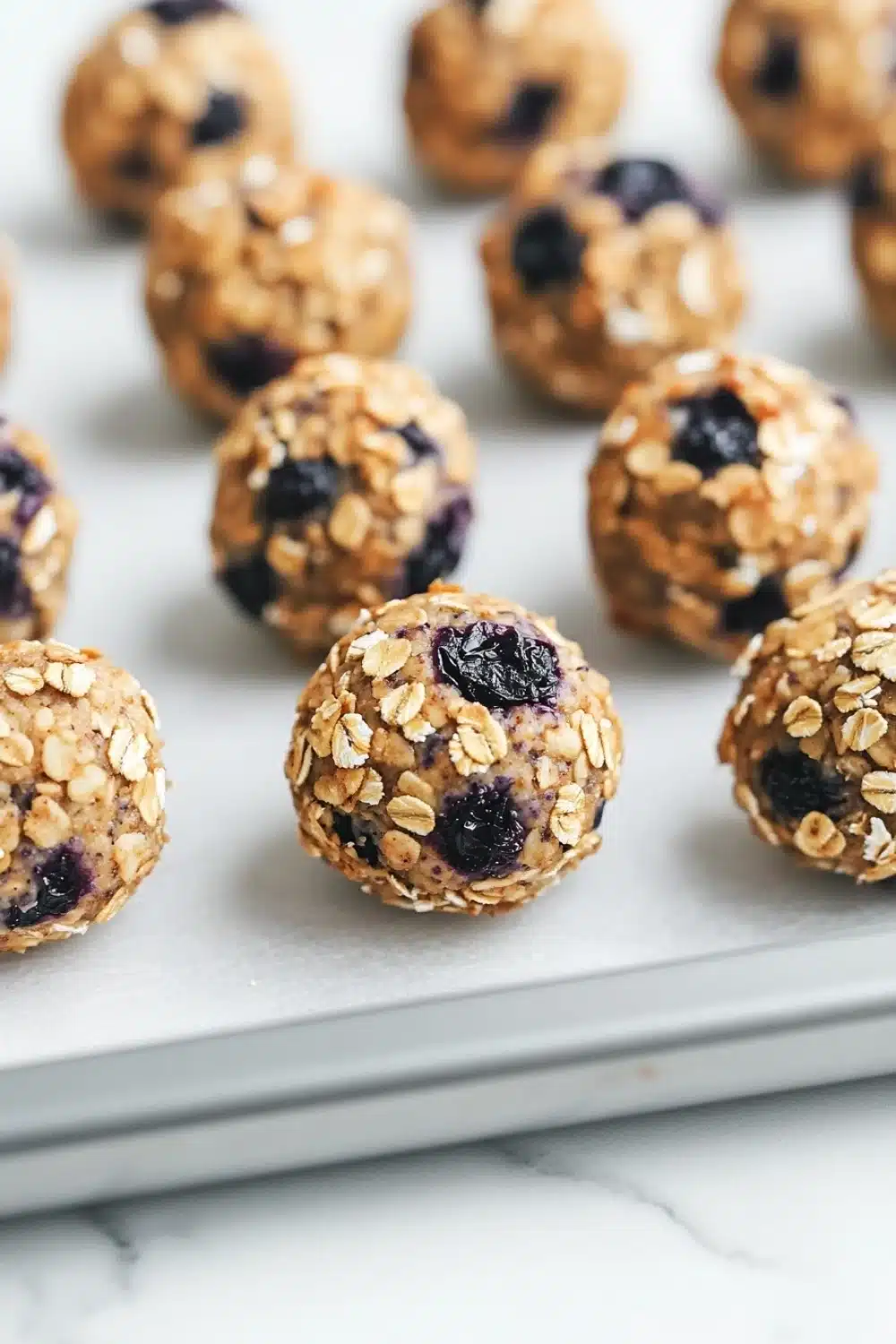 Blueberry muffin protein balls - the image shows a tray of freshly baked granola balls on a white marble countertop. the balls are round and golden brown in color, with visible oats and blueberries scattered throughout. they are arranged in a scattered manner, with some overlapping each other. the tray appears to be lined with parchment paper, and the background is blurred, making the balls the focal point of the image.