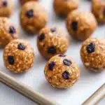 Blueberry muffin protein balls - the image shows a baking tray lined with parchment paper. on the tray, there are round, golden-brown balls that appear to be energy bites. they are covered in sesame seeds and raisins, giving them a crunchy texture. the tray is placed on a white marble countertop. a pair of chopsticks is resting on the tray next to the tray.