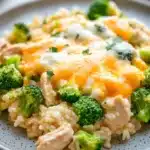 Cheesy chicken broccoli rice casserole one pot - the image shows a plate of food with a serving of rice and broccoli. the rice is white and fluffy, and the broccoli is bright green. on top of the rice, there is a layer of orange-colored cheese, which appears to be melted and bubbly. the cheese is spread evenly over the top, and there are small pieces of chicken scattered throughout the dish. the plate is gray with a speckled pattern, and it is sitting on a wooden table.