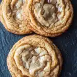 Chewy honey cookie - the image shows three freshly baked cookies on a black slate surface. the cookies are round and golden brown in color, with a crumbly texture on top. they are arranged in a triangular formation, with the largest cookie in the center and two smaller cookies on either side. all three cookies have a swirl pattern on top, with some of the cookies overlapping each other. the background is blurred, making the cookies the focal point of the image.