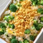 Chicken and broccoli casserole - the image shows a white rectangular baking dish filled with a casserole dish. the dish appears to be made with broccoli, chicken, and cheese. the broccoli is cut into small pieces and is arranged in a circular pattern around the edges of the dish. on top of the broccoli, there is a layer of crumbled breadcrumbs, which are golden brown and appear to be seasoned with herbs and spices. the cheese is melted and bubbly, and there are small pieces of onions scattered throughout the dish, adding a pop of color to the overall appearance. the baking dish is sitting on a wooden table.