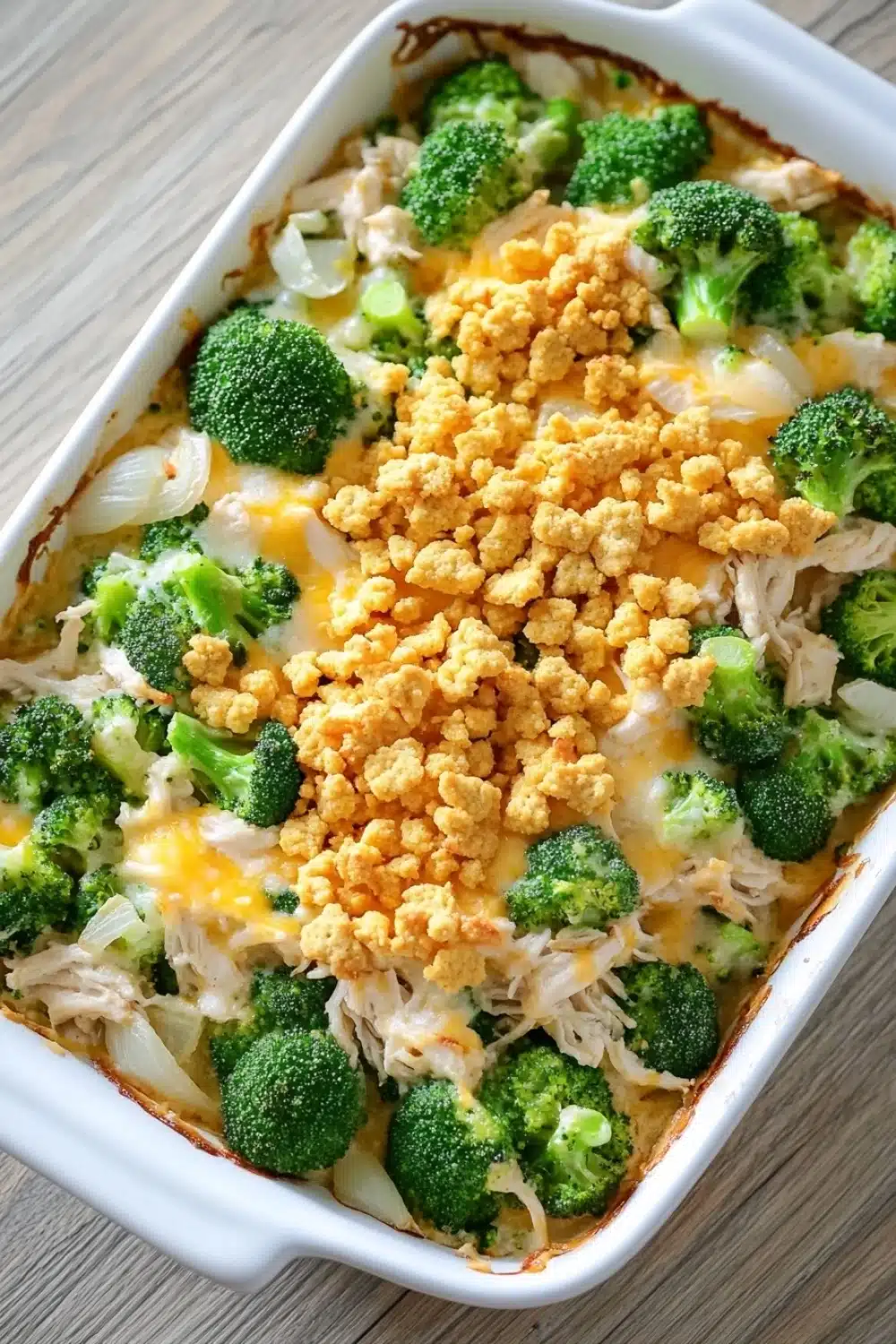 Chicken and broccoli casserole - the image shows a white rectangular baking dish filled with a casserole dish. the dish appears to be made with broccoli, chicken, and cheese. the broccoli is cut into small pieces and is arranged in a circular pattern around the edges of the dish. on top of the broccoli, there is a layer of crumbled breadcrumbs, which are golden brown and appear to be seasoned with herbs and spices. the cheese is melted and bubbly, and there are small pieces of onions scattered throughout the dish, adding a pop of color to the overall appearance. the baking dish is sitting on a wooden table.