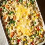 Chicken and rice with veggies casserole oven - the image shows a white rectangular baking dish filled with a casserole dish. the dish appears to be made with rice, vegetables, and chicken, and is topped with melted cheese. the rice is white and fluffy, and the vegetables include carrots, peas, and corn. the cheese is melted and bubbly, and it looks like it has been cooked until it is golden brown. the baking dish is sitting on a wooden table.