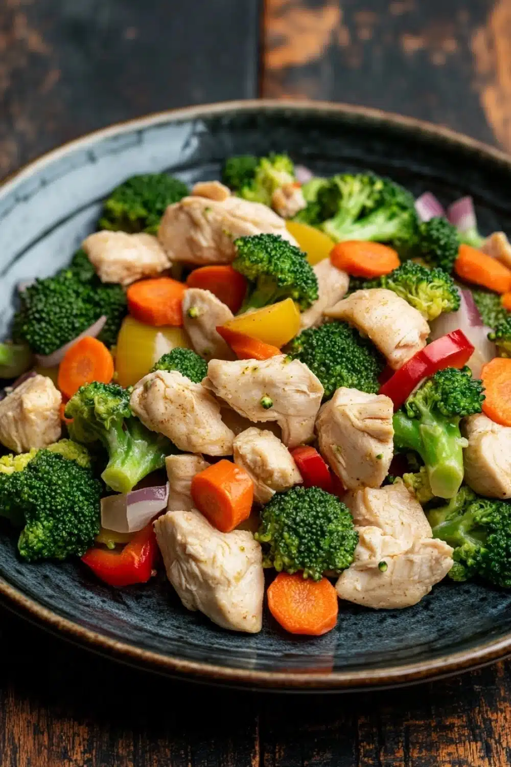 Chicken and vegetables freezer meal - the image is a close-up of a plate of stir-fry with chicken and vegetables. the plate is black and has a textured surface. the chicken is cut into small pieces and is mixed with various vegetables such as broccoli, carrots, bell peppers, and onions. the vegetables are bright green and look fresh and vibrant. the dish is served on a wooden table with a rustic background.