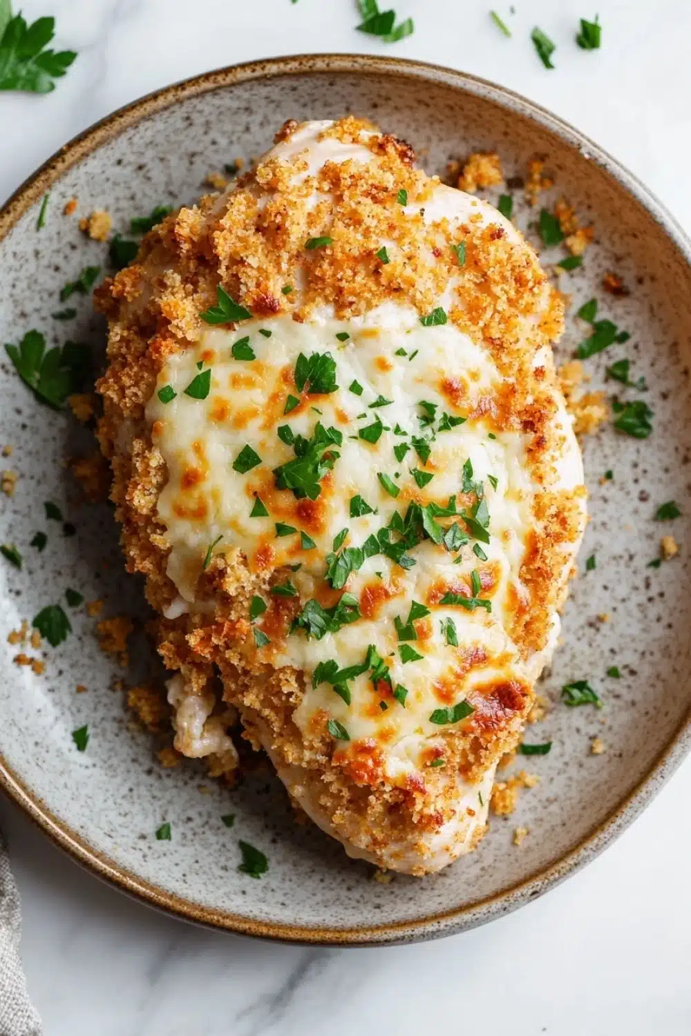 Chicken breast bake - the image shows a plate of food, specifically a piece of breaded chicken. the chicken is golden brown and appears to be seasoned with herbs and spices. the top of the chicken is covered in a layer of melted cheese, which is melted and bubbly. the plate is garnished with chopped parsley, adding a pop of green color to the dish. the background is a white marble countertop with a gray napkin on the side.