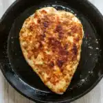 Chicken breast in cast iron skillet - the image shows a piece of cooked chicken in a black cast iron skillet. the chicken appears to be seasoned with herbs and spices, giving it a golden brown color. the skillet is sitting on a white wooden surface, and the chicken is glistening with oil. the word "premium" is visible on the side of the skillet.