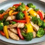 Chicken breast stir fry - the image is a close-up of a plate of stir-fry with chicken and vegetables. the plate is blue and round, and the vegetables are arranged in a colorful and appetizing manner. the chicken is cut into small pieces and appears to be seasoned with herbs and spices. the vegetables include broccoli, red and yellow bell peppers, and green onions. the dish is garnished with a sprinkle of sesame seeds. the background is a wooden table, and there is a watermark in the bottom right corner of the image.