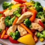 Chicken breast stir fry - the image is a close-up of a plate of stir-fry with chicken and vegetables. the plate is round and has a light brown color with a speckled pattern. the chicken is cut into small pieces and is mixed with various vegetables such as broccoli, red and yellow bell peppers, and orange carrots. the vegetables are cut into thin strips and are arranged in a colorful and appetizing manner. the dish appears to be freshly cooked and ready to eat.
