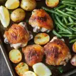 Chicken dinner sheet pan - the image shows a tray of roasted chicken and green beans. the chicken is golden brown and appears to be seasoned with herbs and spices. the green beans are bright green and look fresh and vibrant. there are also small potatoes scattered around the tray. a few lemon wedges are placed on top of the chicken, adding a pop of color to the dish. the tray is lined with parchment paper and is placed on a wooden table.