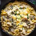 Chicken noodle casserole skillet - the image shows a black cast iron skillet filled with a creamy pasta dish. the dish appears to be a type of fettuccine alfredo, with chunks of chicken and mushrooms mixed in. the pasta is cooked al dente and is topped with melted cheese and chopped parsley. the skillet is sitting on a wooden table with a wooden cutting board and a glass of water in the background. there is also a small bowl of grated cheese on the table. the overall color scheme of the image is warm and inviting.