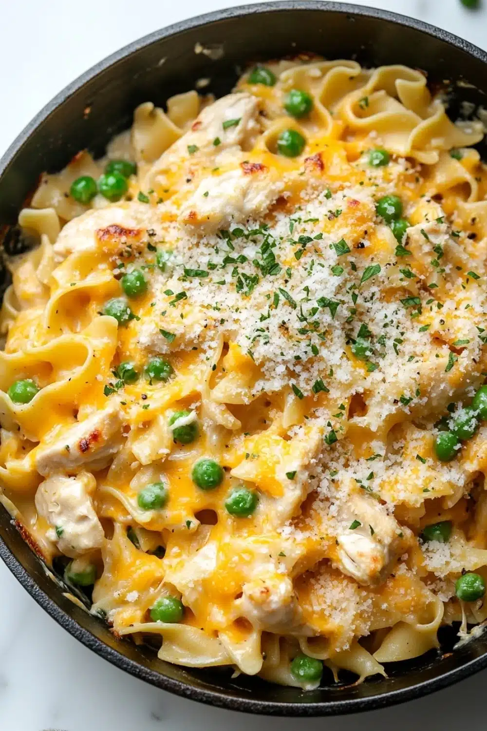Chicken noodle casserole skillet - the image shows a black skillet filled with a creamy pasta dish. the pasta appears to be fettuccine alfredo, with chunks of chicken and peas mixed in. the dish is topped with a layer of melted cheese and sprinkled with grated parmesan cheese. the skillet is sitting on a white marble countertop.