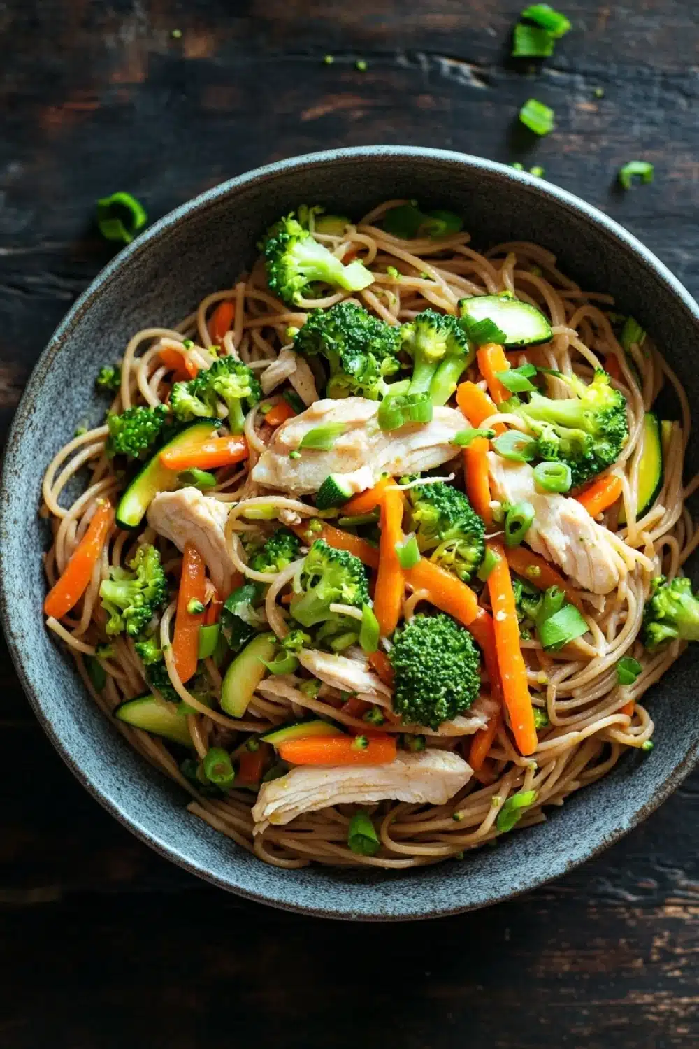 Chicken noodles low calorie - the image is a close-up of a bowl of noodles with chicken and vegetables. the bowl is made of dark blue ceramic and is placed on a dark wooden table. the noodles are light brown and appear to be stir-fried with chicken, broccoli, carrots, and zucchini. the vegetables are bright green and look fresh and vibrant. there are also some chopped green onions scattered throughout the dish. the overall appearance of the dish is fresh and appetizing.