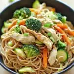 Chicken noodles low calorie - the image is a close-up of a bowl of noodles with chicken and vegetables. the noodles are light brown and appear to be stir-fry. the chicken is cut into small pieces and is mixed with sliced carrots, broccoli, and zucchini. the vegetables are bright green and look fresh and vibrant. the bowl is black and is sitting on a wooden table. there are a few sprigs of green onions scattered around the bowl.