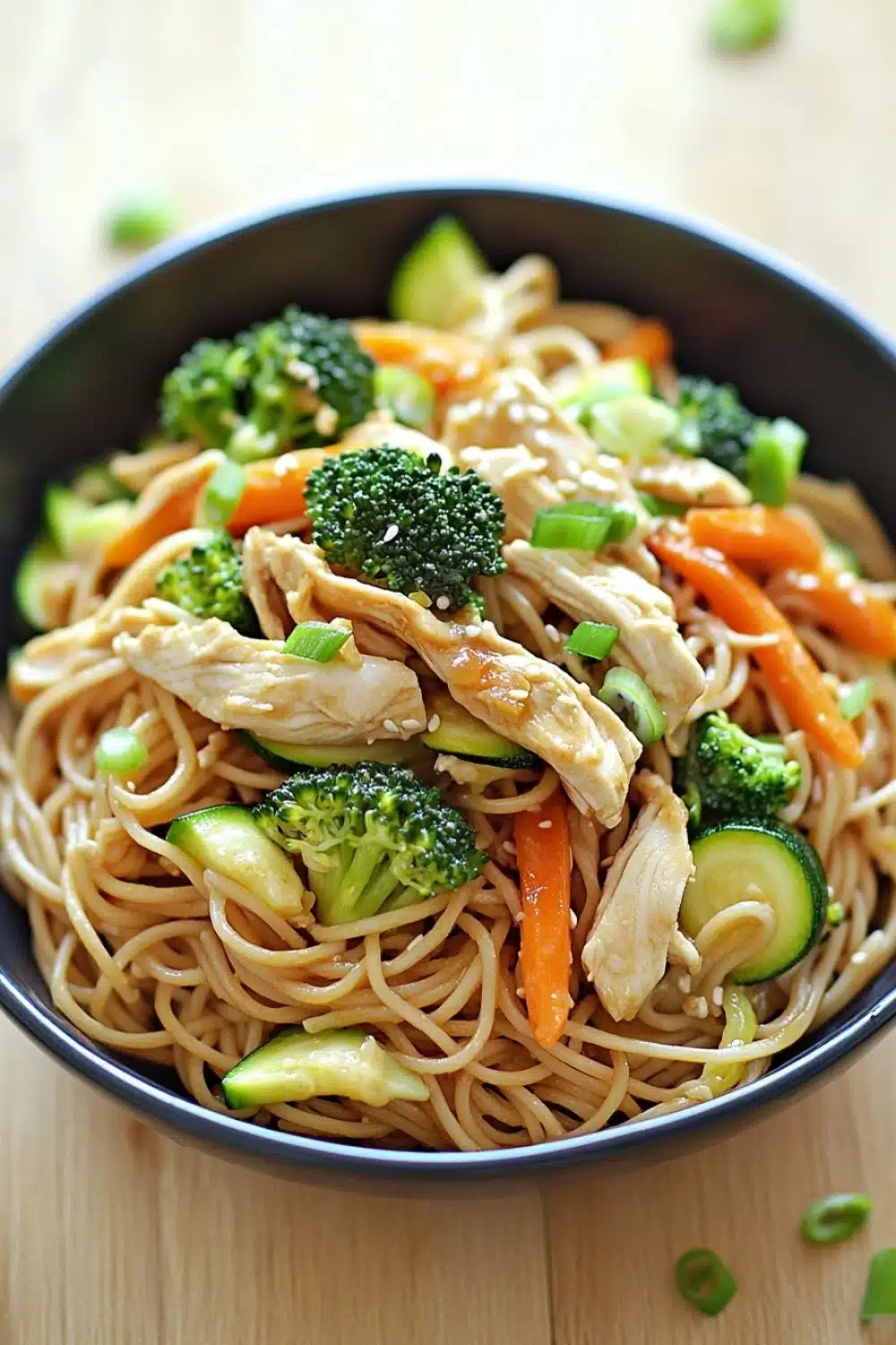 Chicken noodles low calorie - the image is a close-up of a bowl of noodles with chicken and vegetables. the noodles are light brown and appear to be stir-fry. the chicken is cut into small pieces and is mixed with sliced carrots, broccoli, and zucchini. the vegetables are bright green and look fresh and vibrant. the bowl is black and is sitting on a wooden table. there are a few sprigs of green onions scattered around the bowl.