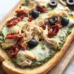 Chicken pesto french bread pizza - the image is of a flatbread pizza on a wooden cutting board. the pizza is topped with shredded chicken, black olives, and sun-dried tomatoes. the chicken is cooked to a golden brown and appears to be seasoned with herbs and spices. the olives are scattered on top of the chicken, adding a pop of color to the dish. the cheese is melted and bubbly, and the tomatoes are bright red. the cutting board is placed on a white marble countertop.