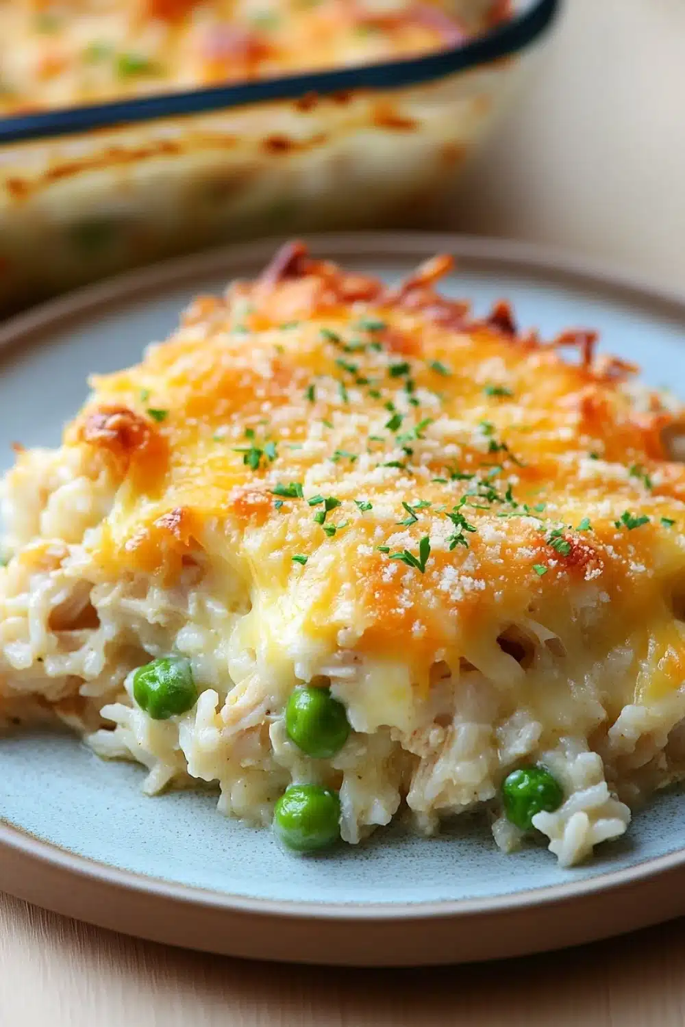 Chicken rice casserole cheese - the image shows a close-up of a dish on a blue plate. the dish appears to be a casserole or macaroni and cheese, with a layer of melted cheese on top. the cheese is golden brown and bubbly, and there are small chunks of green peas scattered throughout the dish. the plate is sitting on a wooden table, and in the background, there is another dish with more of the same dish visible.