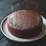 Chocolate cake no icing - the image is a close-up of a round chocolate cake on a white plate. the cake appears to be freshly baked and has a smooth, shiny surface. the plate is placed on a wooden table with a rustic, weathered look. the background is blurred, making the cake the focal point of the image. the focus is on the cake, which is the only object in the frame.