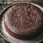 Chocolate cake no icing - the image is a close-up of a round chocolate cake on a white plate. the cake appears to be freshly baked and has a smooth, shiny surface. the plate is placed on a wooden table with a rustic, weathered appearance. the background is blurred, making the cake the focal point of the image. the focus is on the cake, which is the only object in the frame.