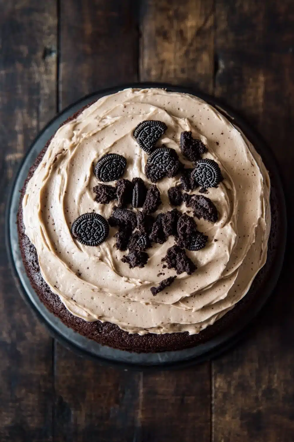 Chocolate cake oreo frosting - the image is of a round chocolate cake on a black cake stand. the cake is covered in a light brown frosting with a swirl pattern on top. on top of the frosting, there are several oreo cookies arranged in a circular pattern. the cookies are dark brown in color and appear to be freshly baked. the background is a wooden table with a rustic texture. the overall mood of the image is warm and inviting.
