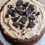 Chocolate cake oreo frosting - the image is of a round chocolate cake on a blue cake stand. the cake is covered in a light brown frosting with a swirl pattern on top. on top of the frosting, there are several oreo cookies arranged in a circular pattern. the cookies are dark brown in color and appear to be freshly baked. the frosting is spread evenly over the cake, creating a smooth and creamy texture. the background is blurred, but it appears to be a wooden table or countertop.