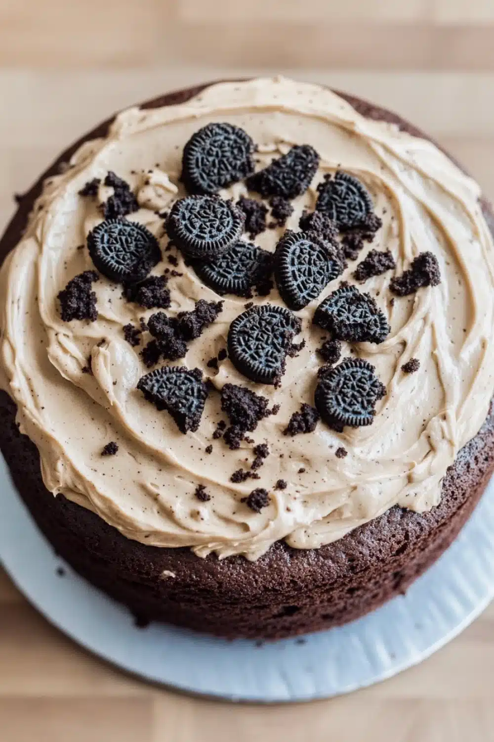 Chocolate cake oreo frosting - the image is of a round chocolate cake on a blue cake stand. the cake is covered in a light brown frosting with a swirl pattern on top. on top of the frosting, there are several oreo cookies arranged in a circular pattern. the cookies are dark brown in color and appear to be freshly baked. the frosting is spread evenly over the cake, creating a smooth and creamy texture. the background is blurred, but it appears to be a wooden table or countertop.