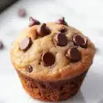 Chocolate chip muffin 4 ingredients - the image shows a freshly baked chocolate chip muffin on a baking tray. the muffin is golden brown in color and has a crumbly texture. it is topped with chocolate chips that are scattered all over the top of the muffin. the tray is lined with parchment paper and there are a few more chocolate chips scattered around the tray.