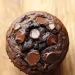 Chocolate muffin cup - the image is a close-up of a round chocolate muffin on a wooden surface. the muffin appears to be freshly baked and has a golden brown color. it is covered in chocolate chips and nuts, giving it a crunchy texture. the chocolate chips are arranged in a circular pattern around the muffin, with some overlapping each other. the background is blurred, making the chocolate chips the focal point of the image.