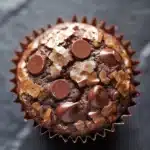 Chocolate muffin cup - the image is a close-up of a chocolate muffin. the muffin is in a paper liner and is placed on a dark grey textured surface. the top of the muffin has a layer of chocolate chips scattered across it, giving it a crunchy texture. there are also several chocolate chips on top, adding a pop of color to the dish. the chocolate chips are arranged in a circular pattern, with some overlapping each other. the overall appearance is decadent and indulgent.