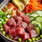 Classic tuna poke bowl - the image is a close-up of a bowl of asian-style salad. the bowl is made of ceramic and has a dark brown color. the salad is made up of various ingredients such as sliced cucumbers, sliced carrots, sliced avocado, sliced green onions, and edamame. there are also sesame seeds sprinkled on top. the ingredients are arranged in a colorful and appetizing manner. the background is dark, making the colors of the ingredients stand out.