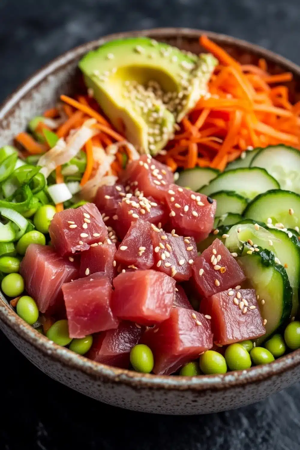 Classic tuna poke bowl - the image is a close-up of a bowl of asian-style salad. the bowl is made of ceramic and has a dark brown color. the salad is made up of various ingredients such as sliced cucumbers, sliced carrots, sliced avocado, sliced green onions, and edamame. there are also sesame seeds sprinkled on top. the ingredients are arranged in a colorful and appetizing manner. the background is dark, making the colors of the ingredients stand out.