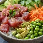Classic tuna poke bowl - the image is a close-up of a bowl of food. the bowl is made of ceramic and has a speckled pattern. inside the bowl, there is a bed of white rice. on top of the rice, there are sliced cucumbers, sliced avocado, and sliced carrots. the vegetables are arranged in a colorful and appetizing manner. there are also some sesame seeds sprinkled on top. the background is blurred, but it appears to be a kitchen countertop with a pair of chopsticks resting on the side.