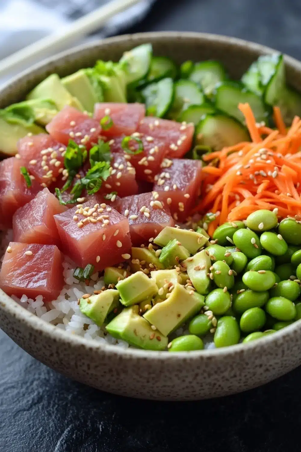 Classic tuna poke bowl - the image is a close-up of a bowl of food. the bowl is made of ceramic and has a speckled pattern. inside the bowl, there is a bed of white rice. on top of the rice, there are sliced cucumbers, sliced avocado, and sliced carrots. the vegetables are arranged in a colorful and appetizing manner. there are also some sesame seeds sprinkled on top. the background is blurred, but it appears to be a kitchen countertop with a pair of chopsticks resting on the side.