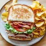 Club sandwich with chips - the image shows a plate with a sandwich and a side of potato chips. the sandwich is made with two slices of toasted bread, filled with a mixture of meat, lettuce, tomato, and bacon. the bread is golden brown and appears to be crispy on the outside. the potato chips are golden and are scattered around the plate. the plate is on a wooden table.