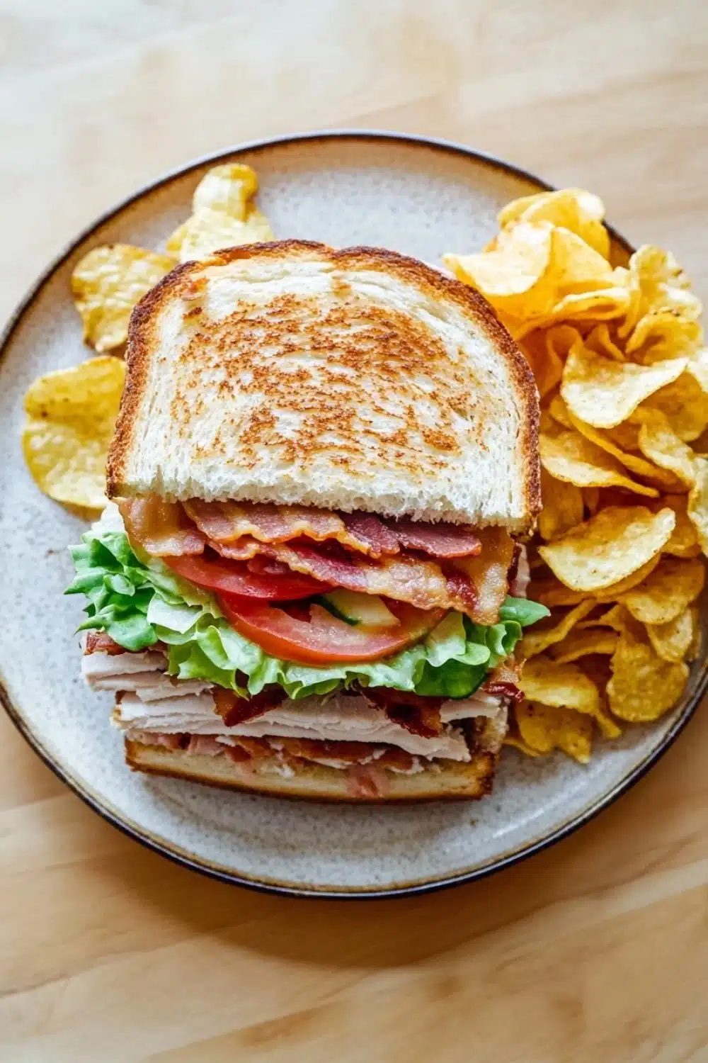 Club sandwich with chips - the image shows a plate with a sandwich and a side of potato chips. the sandwich is made with two slices of toasted bread, filled with a mixture of meat, lettuce, tomato, and bacon. the bread is golden brown and appears to be crispy on the outside. the potato chips are golden and are scattered around the plate. the plate is on a wooden table.