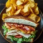 Club sandwich with chips - the image shows a sandwich on a black plate with a pile of potato chips on the side. the sandwich is made with two slices of toasted bread, filled with lettuce, tomato, and bacon. the bacon is crispy and golden brown, and the lettuce is a vibrant green. the plate is sitting on a wooden table with a rustic background.