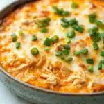 Crock pot buffalo chicken dip - the image shows a close-up of a casserole dish with a creamy orange-colored dip in it. the dip appears to be made with shredded chicken, cheese, and chopped green onions. the dish is sitting on a white marble countertop. the edges of the dish are slightly frayed, indicating that it has been cooked. the overall appearance is appetizing and ready to eat.
