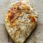 Crock pot chicken breast - the image shows a piece of grilled chicken breast on a wooden cutting board. the chicken breast is golden brown and appears to be seasoned with herbs and spices. the skin of the chicken is crispy and golden brown, indicating that it has been cooked to perfection. the seasoning is visible on the top of the breast, with small flecks of herbs scattered throughout. the cutting board has a light-colored wood grain pattern, and the background is blurred, making the chicken the focal point of the image.