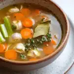 Crock pot chicken broth - the image is a close-up of a bowl of soup. the soup appears to be a vegetable soup with chunks of carrots, celery, onions, and other vegetables mixed in. there is a sprig of thyme on top of the soup, adding a pop of green color to the dish. the bowl is sitting on a white plate with a speckled pattern. the background is blurred, making the soup the focal point of the image.